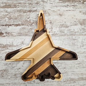 May include: Wooden airplane-shaped tray with a layered design. The tray features a mix of light and dark wood tones, including shades of brown and tan, arranged in diagonal stripes. The tray is designed to resemble a fighter jet.