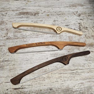 May include: Three wooden cheese slicers with metal wires. The top slicer is light wood with an engraved letter A. The middle slicer is medium-toned wood with the text Tessa's Kitchen engraved. The bottom slicer is dark wood with a letter J engraved.