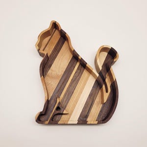 May include: A cat-shaped wooden tray constructed from alternating light and dark wood strips. The tray features a raised border and a silhouette design of a sitting cat with a curled tail, showcasing natural wood grain patterns throughout the piece.