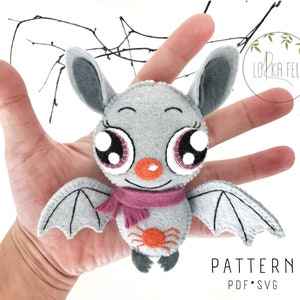K&ouml;nnte beinhalten: Eine graue Filzfledermaus mit gro&szlig;en rosa Augen, einem rosa Schal und einer orangefarbenen Spinne auf der Brust. Die Fledermaus wird von einer Hand gehalten. Der Text "PATTERN PDF * SVG" befindet sich am unteren Rand des Bildes.