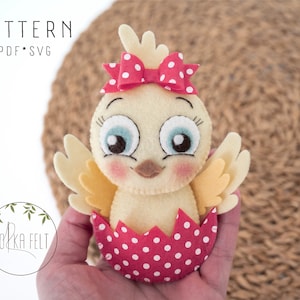 Op de afbeelding: Een geel vilt kuiken met een rode strik met witte stippen, zittend in een rode eierschaal met witte stippen. Het kuiken heeft blauwe ogen en een roze blos.