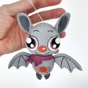 K&ouml;nnte beinhalten: Eine graue Filzfledermaus mit gro&szlig;en Augen, einem rosa Schal und einer roten Spinne auf der Brust. Die Fledermaus h&auml;ngt an einer wei&szlig;en Schnur.