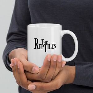 Op de afbeelding: Een witte keramische mok met een zwarte afbeelding met de tekst "The Reptiles".