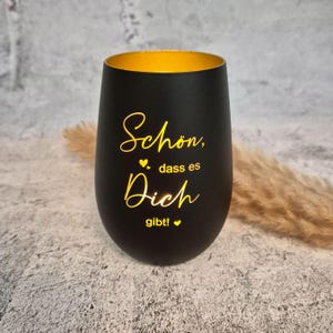 May include: A black stemless wine glass with a gold interior. The glass has the words "Sch&ouml;n, dass es Dich gibt!" in gold script. The glass is on a textured surface.