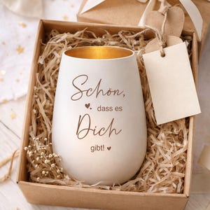 May include: A white, egg-shaped candle holder with gold interior, inscribed with the words "Sch&ouml;n, dass es Dich gibt!" in brown script. It rests in a brown box with shredded paper, a gift box, and a blank tag.