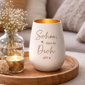 May include: A white ceramic candle holder with gold interior and the text "Sch&ouml;n, dass es Dich gibt!" in brown script. A lit tea light and a small vase of flowers sit on a wooden tray, creating a cozy atmosphere.