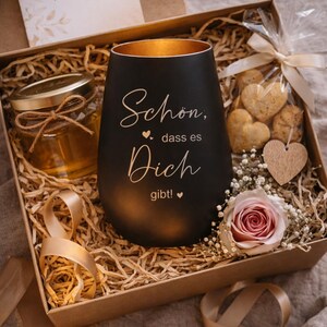May include: A gift box with a black candle holder, a jar of honey, heart-shaped cookies, and a rose. The candle holder has gold interior and the words "Sch&ouml;n, dass es Dich gibt!" The box includes decorative straw and a card that says "F&uuml;r Dich".