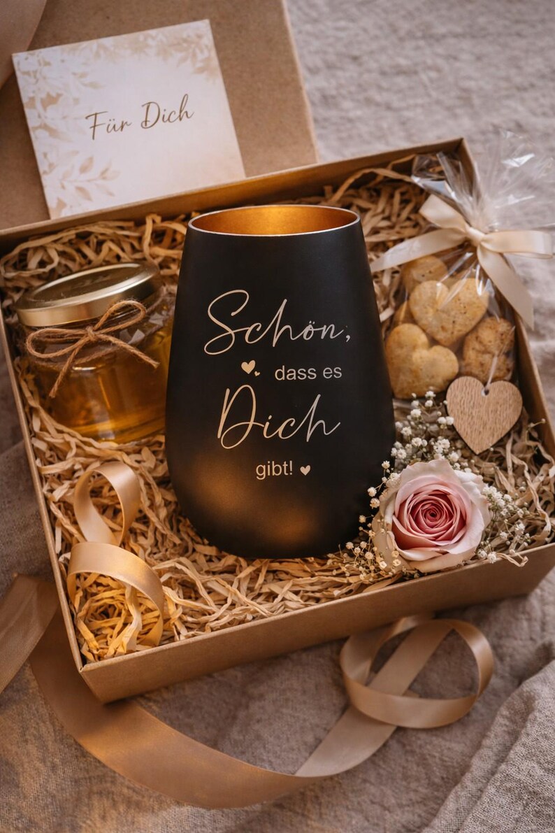 May include: A gift box with a black candle holder, a jar of honey, heart-shaped cookies, and a rose. The candle holder has gold interior and the words "Sch&ouml;n, dass es Dich gibt!" The box includes decorative straw and a card that says "F&uuml;r Dich".