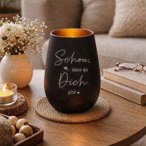 May include: A black candle holder with a gold interior, featuring the text "Sch&ouml;n, dass es Dich gibt!" in white script. The candle holder sits on a woven coaster on a wooden table, with a vase of white flowers in the background.