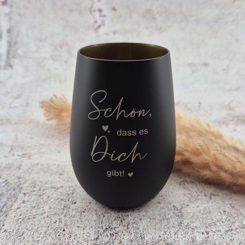 May include: A black stemless wine glass with gold interior. The glass has the words "Sch&ouml;n, dass es Dich gibt!" in a white script font. A small heart symbol is between the words. The glass is on a light gray surface.