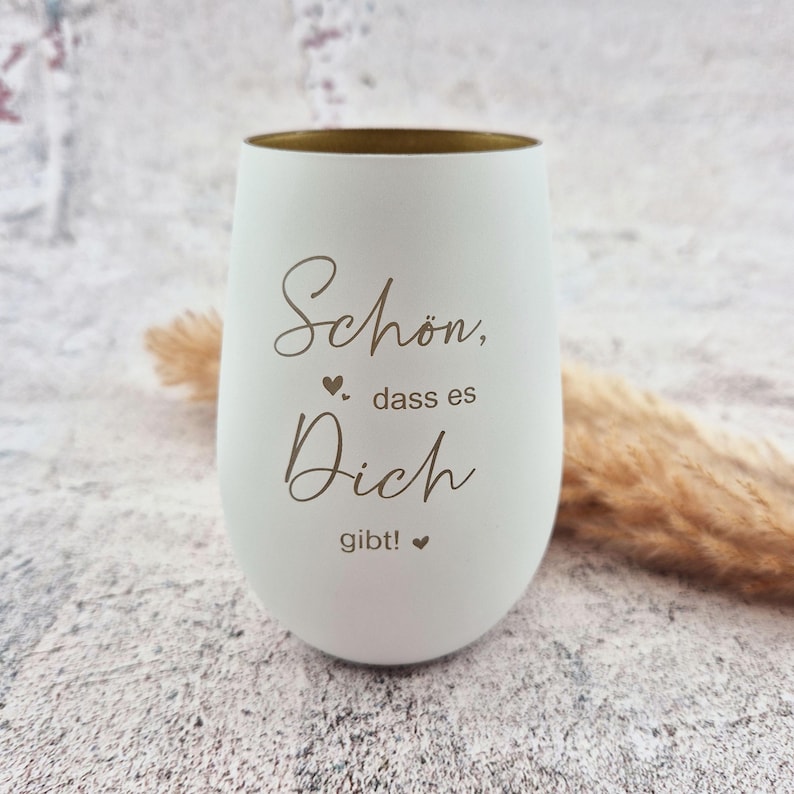 May include: A white stemless wine glass with a gold rim. The glass has the words "Sch&ouml;n, dass es Dich gibt!" etched in a script font, with heart symbols. The glass is on a neutral background.