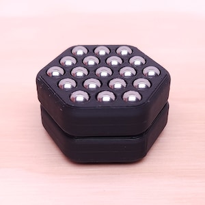 May include: A black hexagonal box with a lid, containing a grid of silver metal balls. The box is made of 3D printed plastic.
