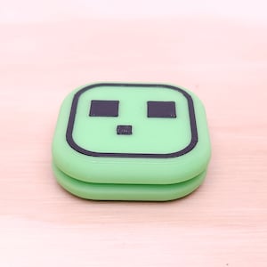 May include: A green and black plastic case with a simple face design. The case has two black eyes and a small mouth.