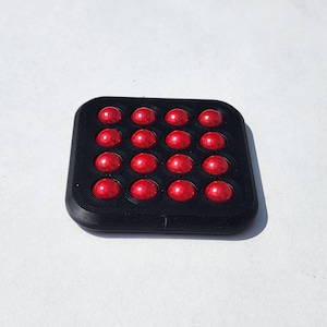 May include: A square black fidget toy featuring a four-by-four grid of sixteen glossy red spherical buttons. The device has rounded corners and a matte black frame that holds each individual red sphere in a recessed circular socket.