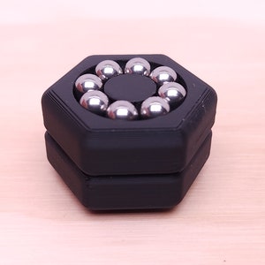 May include: A black hexagonal fidget toy with a circular groove containing eight silver metal balls.
