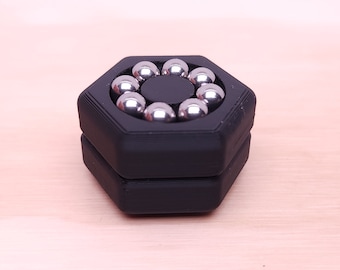 Puede incluir: Un juguete de fidget hexagonal negro con una ranura circular que contiene ocho bolas de metal plateadas.