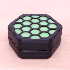 May include: A black hexagonal container with a green honeycomb pattern on the top. The container is made of plastic and has a smooth surface.