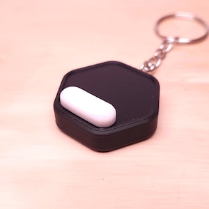 May include: A black, hexagonal keychain with a white, pill-shaped object on top. The keychain has a silver chain and ring. The items are on a light brown wooden surface.