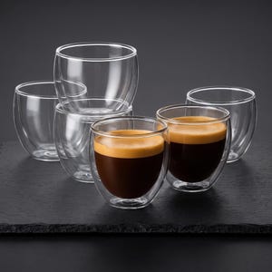 May include: A set of six clear double-walled glass cups. Two of the cups are filled with dark brown espresso topped with a light brown crema, while the remaining four cups are empty. The glasses feature a rounded, handleless design.