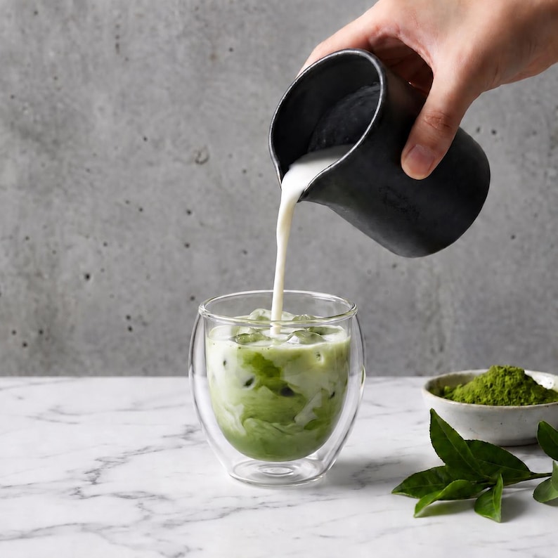 May include: A clear double-walled glass containing a green matcha latte with ice cubes. A matte black ceramic pitcher pours white milk into the glass. A small bowl filled with green matcha powder and several green tea leaves sit nearby.