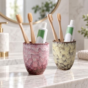 May include: Two ceramic tumblers with a textured, mottled glaze in shades of pink and yellow. Each tumbler holds two bamboo toothbrushes and a white tube of toothpaste with a green label. The containers have a rounded shape and a speckled finish.