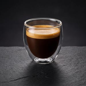 May include: A clear double-walled glass cup filled with dark brown espresso topped with a light brown crema. The vessel features a rounded base and a smooth rim, showcasing the liquid contents through the transparent glass walls.