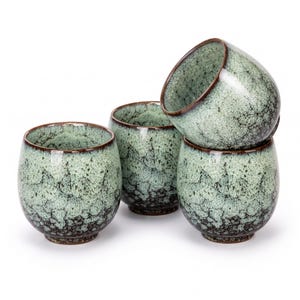 May include: A set of four ceramic cups featuring a speckled light green and dark brown reactive glaze. The cups have a rounded, bulbous shape with a smooth rim and a dark brown base, showcasing a distinct crackle pattern across the exterior surface.