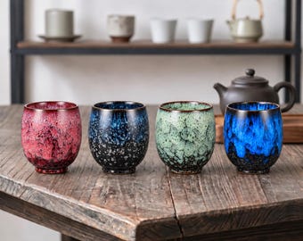 May include: Four ceramic tea cups with a rounded shape and a reactive glaze finish. The cups feature distinct color gradients: one in deep red, one in dark blue and black, one in light green, and one in a vibrant blue and black speckled pattern.