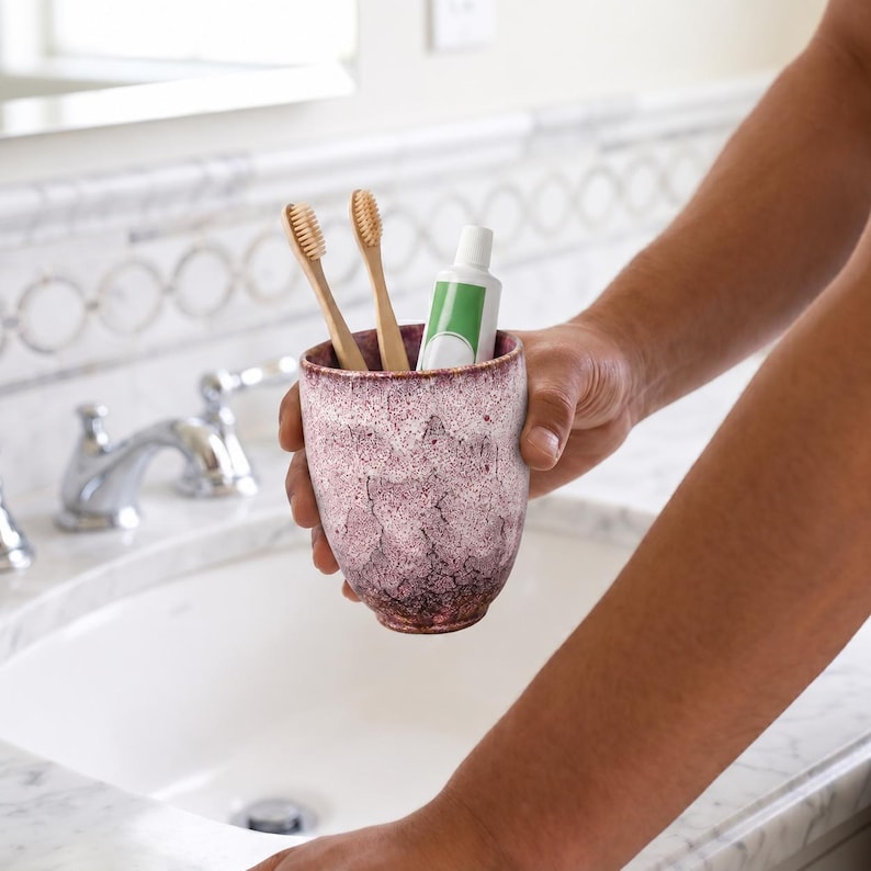 Pode incluir: Um copo de cer&acirc;mica com acabamento salpicado em tons de vermelho escuro e branco, contendo duas escovas de dentes de bambu e um tubo de pasta de dentes branco com um r&oacute;tulo verde. O recipiente tem uma forma arredondada e texturizada.