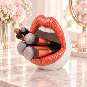Pode incluir: Um suporte de cerâmica branca em forma de boca aberta com lábios vermelhos brilhantes e dentes brancos, contendo vários pincéis de maquilhagem com cabos cor de cobre e cerdas macias e matizadas para organizar ferramentas cosméticas.