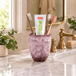 Pode incluir: Um copo de cer&acirc;mica texturizado, com manchas roxas e brancas, cont&eacute;m um tubo de pasta de dentes branca com o r&oacute;tulo Brand Fresh Mint e tr&ecirc;s escovas de dentes com cerdas brancas, castanho-claras e cor-de-rosa na vertical.