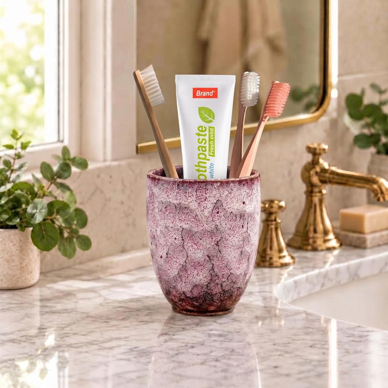 Pode incluir: Um copo de cer&acirc;mica texturizado, com manchas roxas e brancas, cont&eacute;m um tubo de pasta de dentes branca com o r&oacute;tulo Brand Fresh Mint e tr&ecirc;s escovas de dentes com cerdas brancas, castanho-claras e cor-de-rosa na vertical.