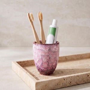 Pode incluir: Um copo cil&iacute;ndrico de cer&acirc;mica com um esmalte texturizado e manchado em rosa e branco, contendo duas escovas de dentes de bambu e um tubo de pasta de dentes branco com um r&oacute;tulo verde. O copo tem uma base c&oacute;nica e um acabamento rugoso.