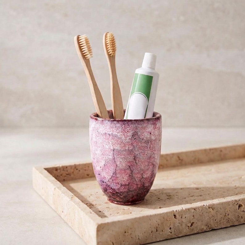 Pode incluir: Um copo cil&iacute;ndrico de cer&acirc;mica com um esmalte texturizado e manchado em rosa e branco, contendo duas escovas de dentes de bambu e um tubo de pasta de dentes branco com um r&oacute;tulo verde. O copo tem uma base c&oacute;nica e um acabamento rugoso.