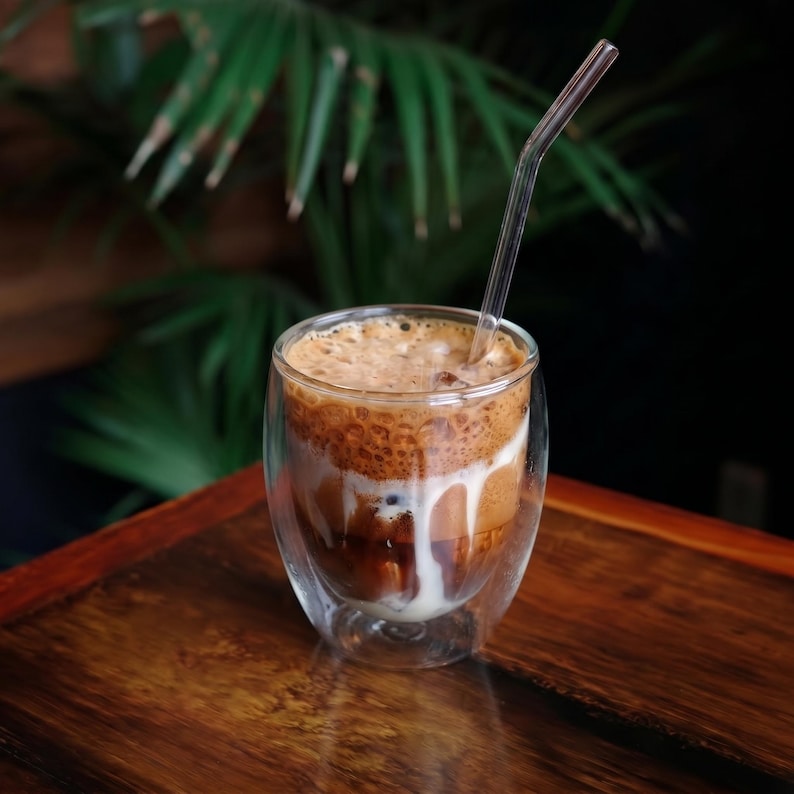 May include: A double-walled glass tumbler filled with an iced coffee beverage featuring a distinct layer of dark coffee, white milk, and a thick, foamy brown top. A clear, bent glass straw is inserted into the drink, extending above the rim.