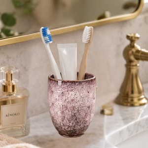 Pode incluir: Um suporte de cer&acirc;mica para escovas de dentes com acabamento texturizado e manchado em tons de rosa antigo e castanho escuro. Dentro do suporte est&atilde;o duas escovas, uma com cabo branco e cerdas azuis, outra com cabo de madeira clara e cerdas brancas.