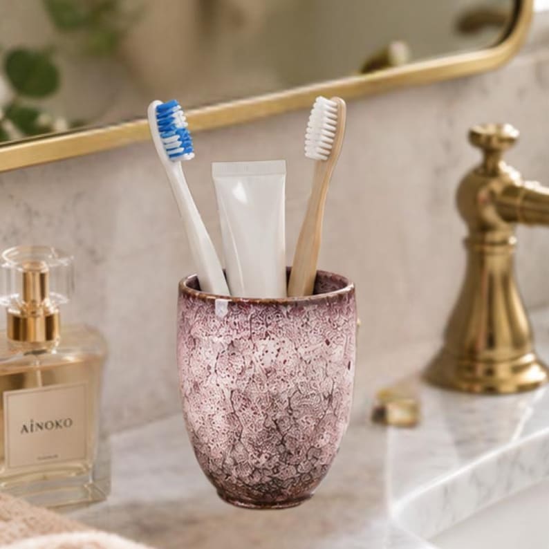 Pode incluir: Um suporte de cer&acirc;mica para escovas de dentes com acabamento texturizado e manchado em tons de rosa antigo e castanho escuro. Dentro do suporte est&atilde;o duas escovas, uma com cabo branco e cerdas azuis, outra com cabo de madeira clara e cerdas brancas.