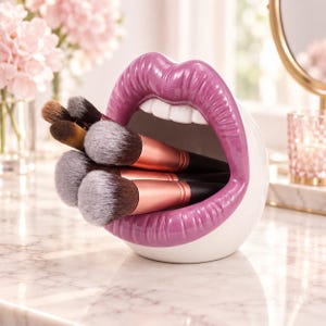 Pode incluir: Um suporte de cerâmica branca para pincéis de maquilhagem em forma de lábios abertos com batom roxo brilhante. O suporte contém vários pincéis com cabos pretos, virolas em ouro rosa e cerdas macias em tons de castanho e branco.