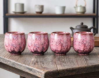 May include: A set of four ceramic tea cups featuring a reactive glaze with a gradient of deep magenta and soft pink tones. Each cup has a rounded, handleless silhouette with a textured, mottled finish that transitions to a darker base.
