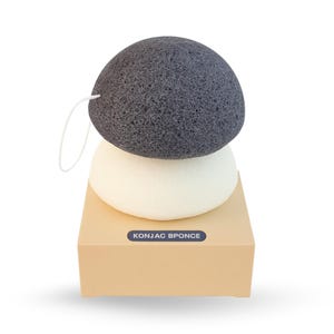 May include: Two konjac sponges, one charcoal gray and one off-white, stacked on a small, light brown box. The gray sponge has a white string loop. The box has the text "KONJAC BPONCE" in white on a blue rectangle.