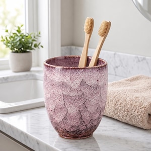 Pode incluir: Um copo cil&iacute;ndrico de cer&acirc;mica com acabamento salpicado e marmoreado em tons de ameixa escuro, magenta e branco, contendo duas escovas de dentes de madeira clara com cerdas macias posicionadas verticalmente no interior.