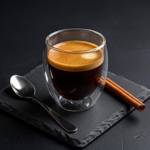 May include: A double-walled glass cup filled with dark brown espresso topped with a golden crema. A silver metal spoon rests to the left of the glass, and two brown cinnamon sticks are positioned to the right on a dark textured surface.