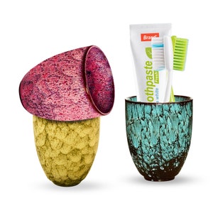 May include: Two ceramic cups, one yellow and one pink, with a turquoise cup holding a toothbrush and a tube of toothpaste that says "Brand" and "toothpaste" with a green leaf design. The toothpaste says "Fresh" and "white" on the tube.