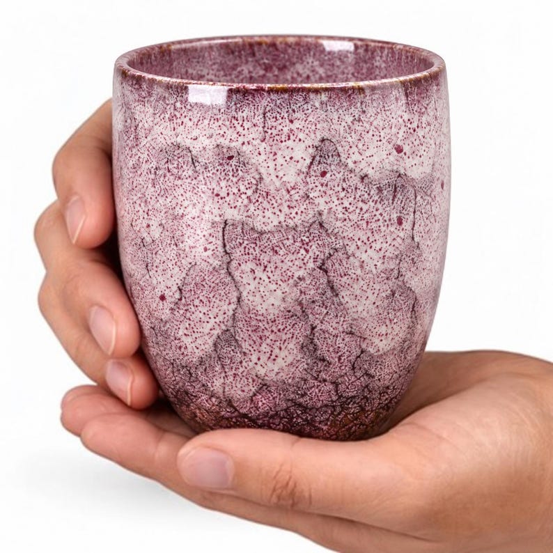 Pode incluir: Uma ch&aacute;vena cil&iacute;ndrica de cer&acirc;mica com um esmalte salpicado em tons de magenta profundo e branco. A superf&iacute;cie apresenta um padr&atilde;o de fissuras distinto, com um rebordo liso e arredondado e uma base c&oacute;nica, exibindo um acabamento texturado.