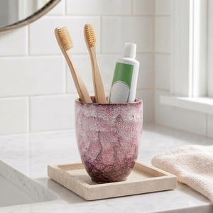 May include: A cylindrical ceramic tumbler with a mottled pink and white glaze finish, containing two bamboo toothbrushes and a white toothpaste tube with a green label, resting on a square beige stone tray with a smooth, flat surface.