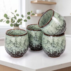 May include: A set of four rounded ceramic cups featuring a reactive glaze with a pale mint green and white speckled finish that transitions into a dark brown base. Each cup has a smooth, curved silhouette and a distinct dark brown rim.