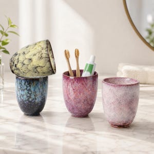 May include: Four ceramic bathroom tumblers in shades of blue, yellow, and pink. One tumbler holds two toothbrushes and a tube of toothpaste. The tumblers have a speckled, textured finish and are arranged on a marble countertop.