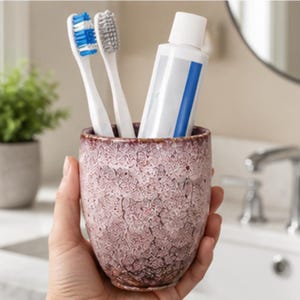 Pode incluir: Um copo cil&iacute;ndrico de cer&acirc;mica com um esmalte texturizado e rachado em tons de rosa empoeirado e castanho escuro. O copo cont&eacute;m duas escovas de dentes, uma com cerdas azuis e brancas e outra com cerdas cinzentas, al&eacute;m de um tubo de pasta de dentes.