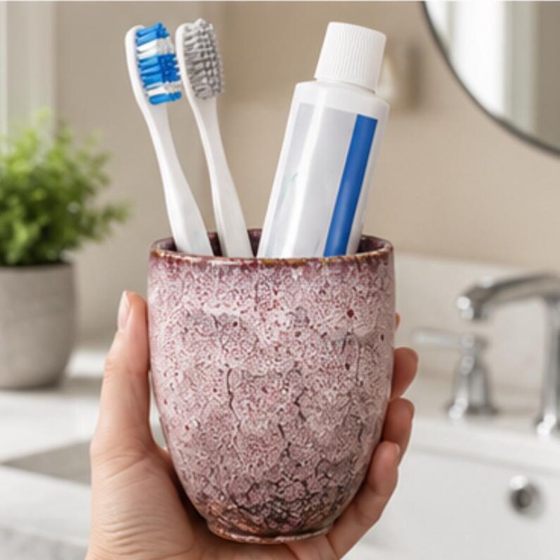 Pode incluir: Um copo cil&iacute;ndrico de cer&acirc;mica com um esmalte texturizado e rachado em tons de rosa empoeirado e castanho escuro. O copo cont&eacute;m duas escovas de dentes, uma com cerdas azuis e brancas e outra com cerdas cinzentas, al&eacute;m de um tubo de pasta de dentes.