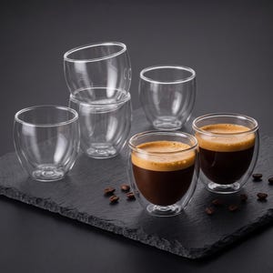 Double wall glass espresso cups filled with fresh espresso, arranged on a dark slate tray with coffee beans.